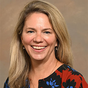 A headshot photo of a woman with long blonde hair, smiling in a blue and red patterned blouse.