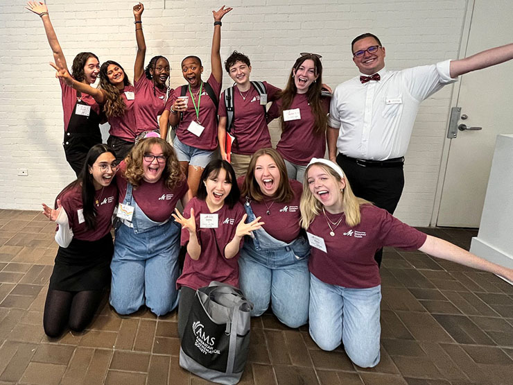 A group of 11 college students in matching maroon T-shirts and one staff member in a white button down and bow tie smile and reach their arms out.