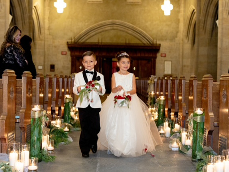 Two children, dressed in a formal suit and fluffy dress and holding rings and a basket of flowers, walk down the aisle of an chapel.