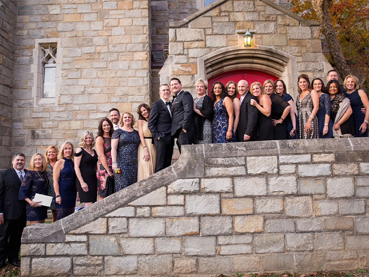 A crowd of wedding guests gather around two newlywed grooms outside of a stone chapel.