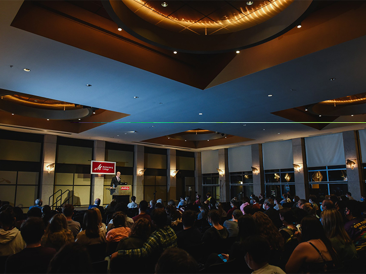 A speaker at a podium addresses a room full of people under dramatic lights.