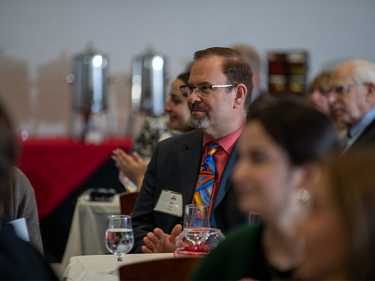 An adult in a suit and tie sits at a table in a room full of formally-dressed adults at tables with white tablecloths.