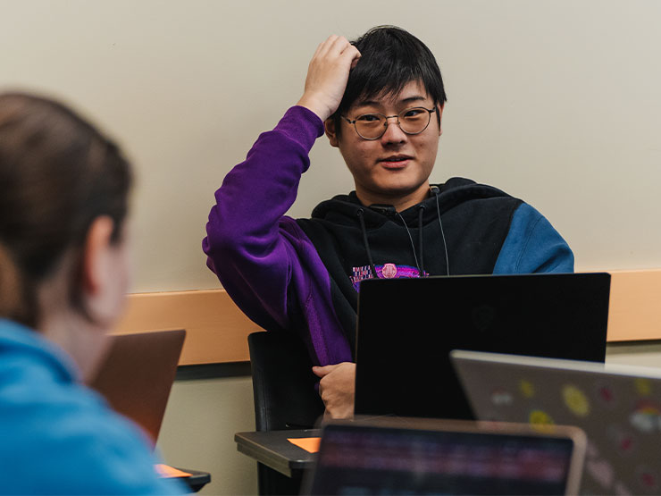 A young adult in a classroom scratches their head while engaged in a discussion.