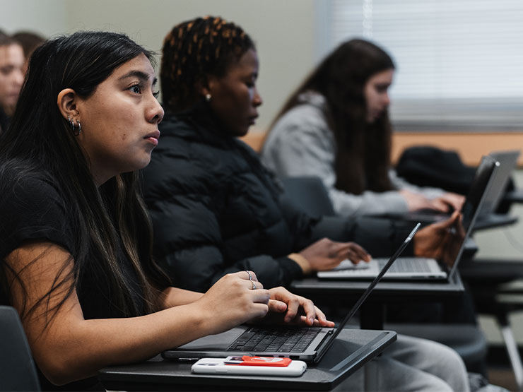 College students listen in a classroom with notebooks and laptops open on their desks.