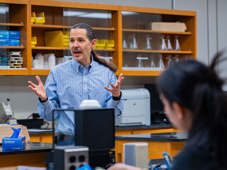 An instructor, with a long ponytail and blue dress shirt, speaks to students in a college laboratory.