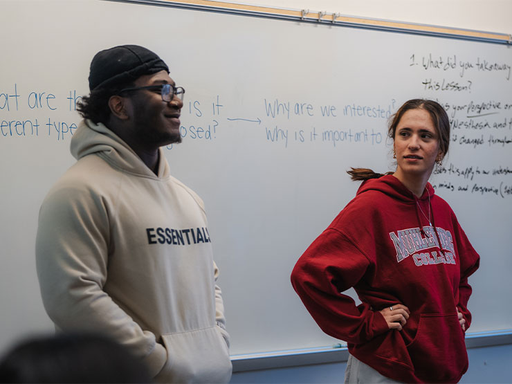 A pair of students stand at the front of the class, a whiteboard with notes behind them.