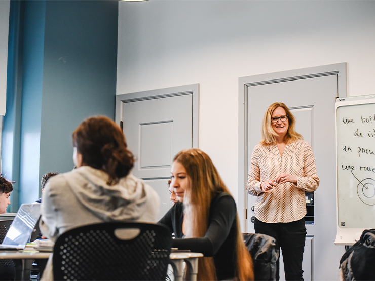 Students sit around small tables while an adult with shoulder-length blonde hair and a a cream-colored blouse addresses them,