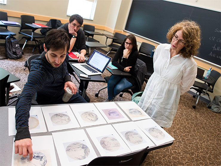 An instructor looks over a rows of Japanese art displayed on a student's desk in a classroom.