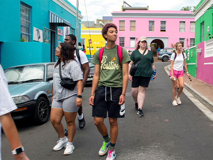 A group of students walk down the streets of a South African neighborhood, dotted with brightly colored blue, yellow, pink and green buildings.