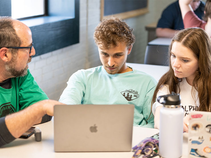 An instructor speaks to two students who looking intently at an open laptop.