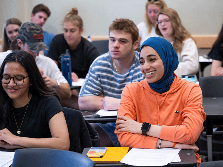 A classroom of students sitting at desks smile at someone off-camera.