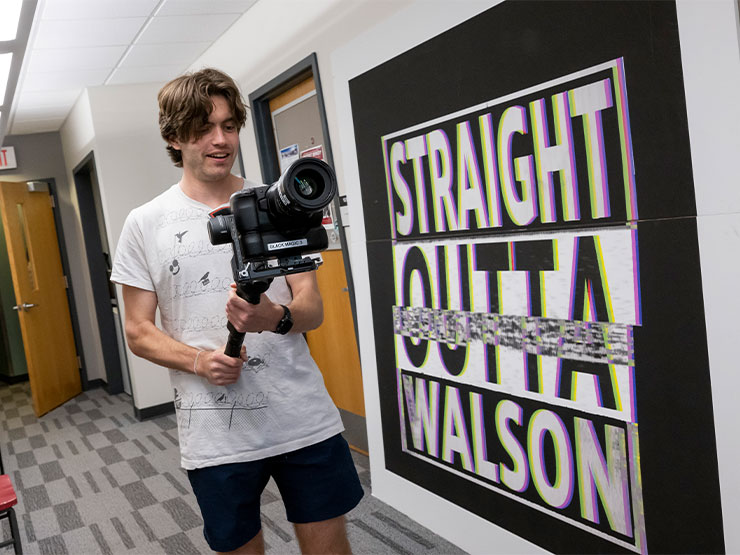 A young adult works on a camera attached to a tripod in a college building that includes a sign that reads 