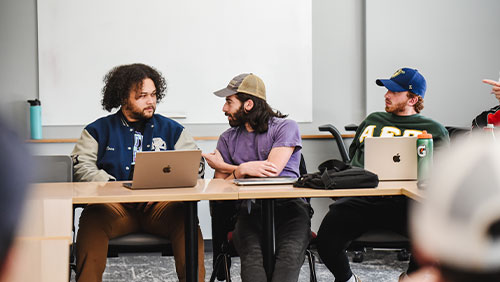 A group students in a classroom have a discussion across a table of open laptops.