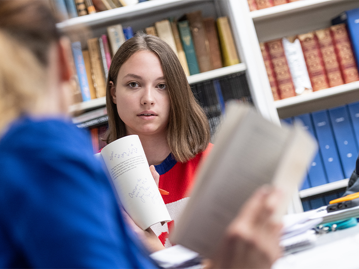 A student holds open a book with notes toward the camera in a room surrounded by reference books.