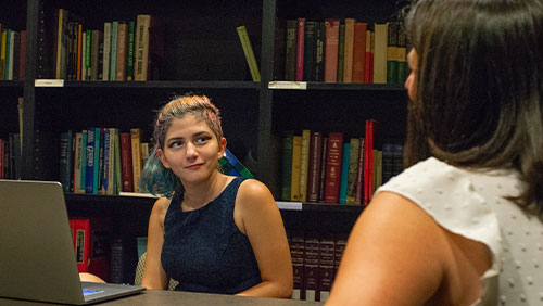 A young adult smiles during a conversation with an adult across a table in a room lined with books.