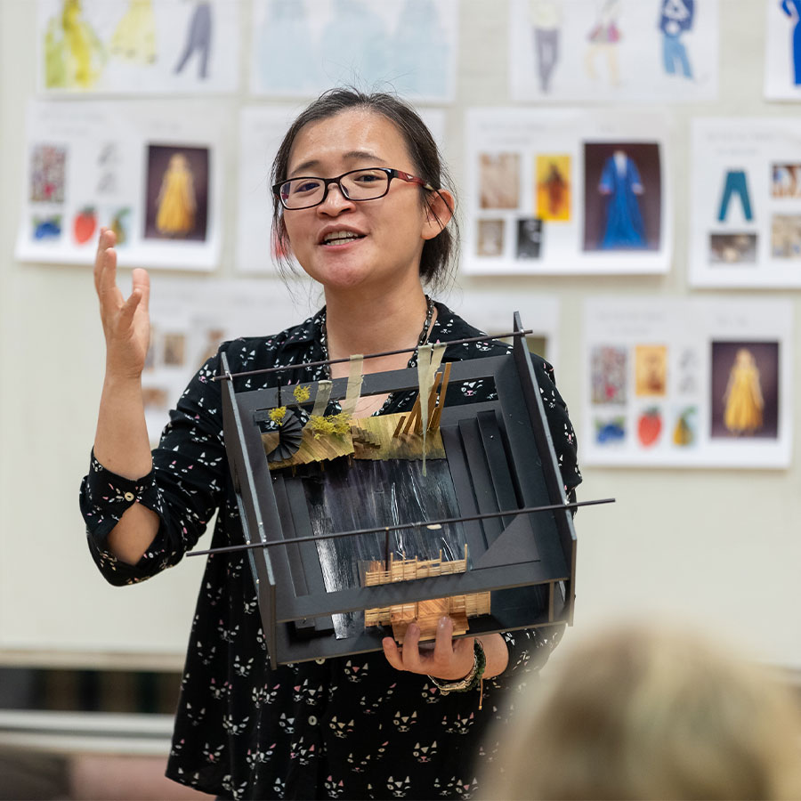 An adult instructor holds a model of a stage, behind them is a whiteboard covered in printed papers showing costume designs.