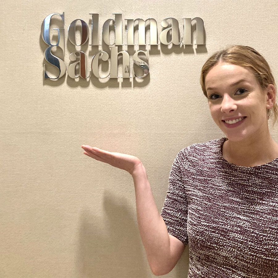 A young adult in a dress shirt smiles and gestures to a sign on the wall that reads Goldman Sachs.