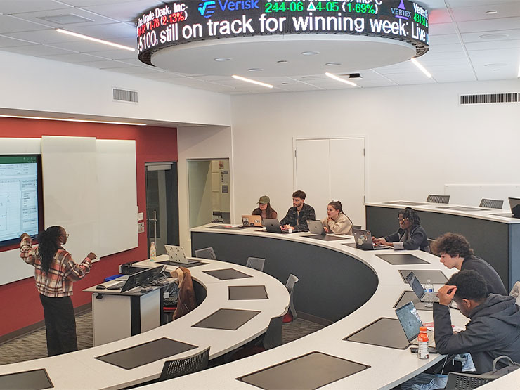 A student stands in front of a classroom, pointing to a projection while students look on, a circular stock ticker suspended on the ceiling above.