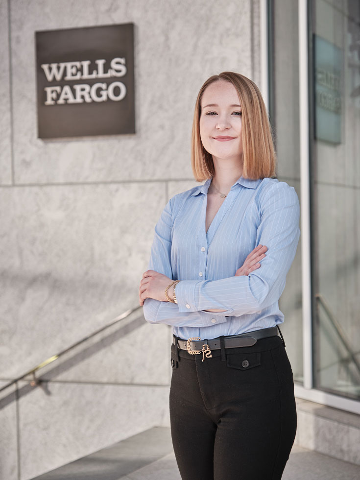 A young adult with shoulder-length blonde hair stands in front of a building entrance that reads