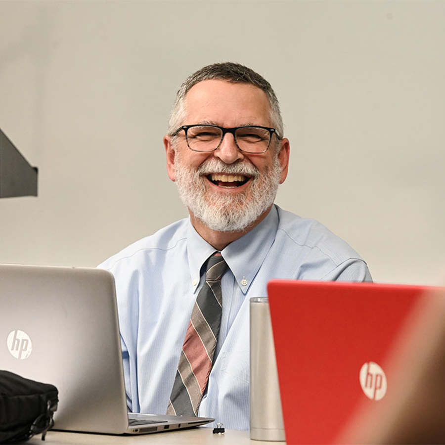 A male instructor, wearing a suit and tie, smiles while seated at a table with open laptops.