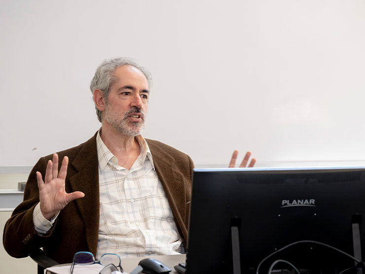 An adult instructor wearing a white dress shirt and dark brown jacket speaks to students off-camera while seated as a table.