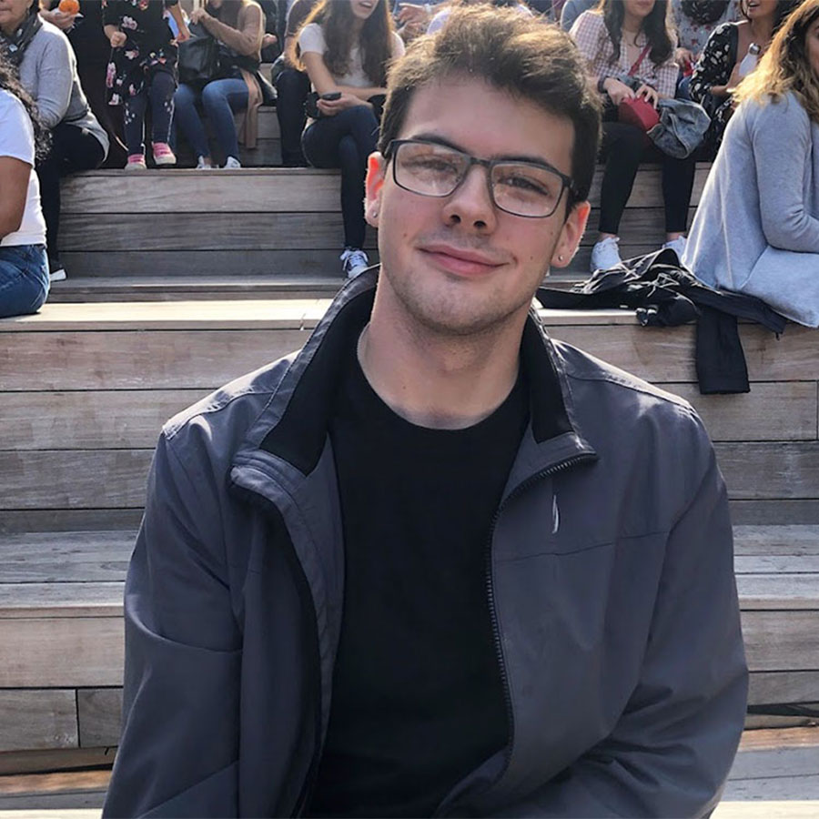 A young adult in a black shirt, grey jacket and glasses smiles softly at the camera while sitting in an outdoor area surrounded by people.