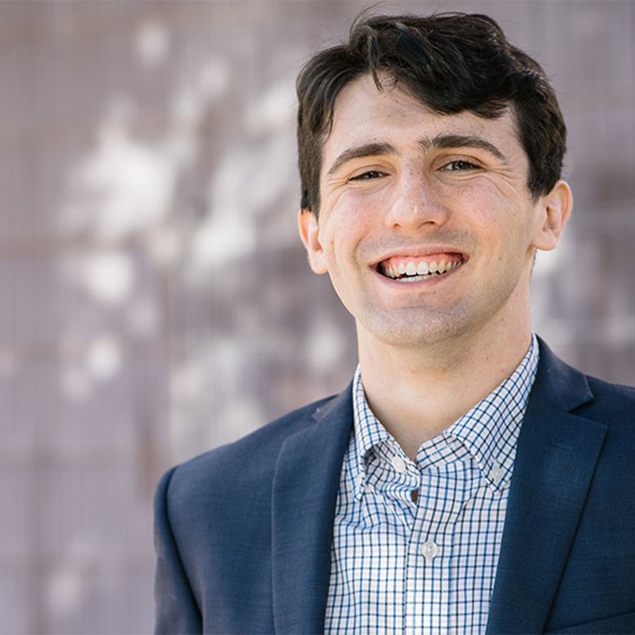 A young adult wearing a dark blue suit jacket and blue and white dress shirt grins at the camera.