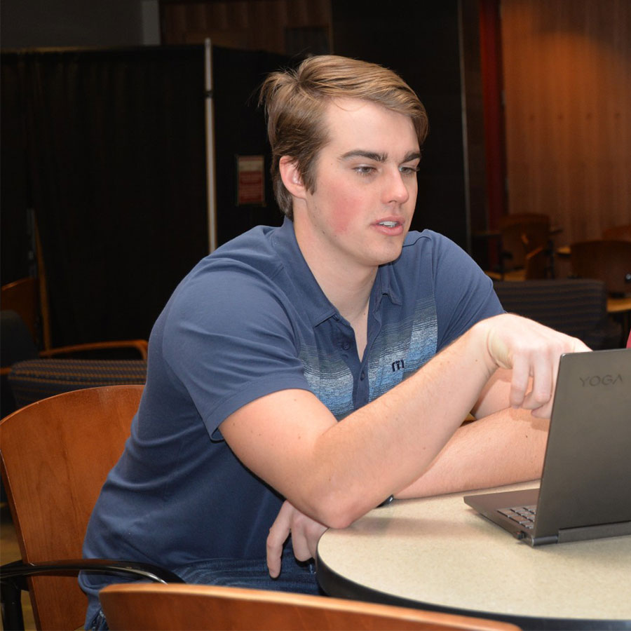 A student in a blue short-sleeved shirt sits at a raised table in a common area and points to the screen of a laptop.