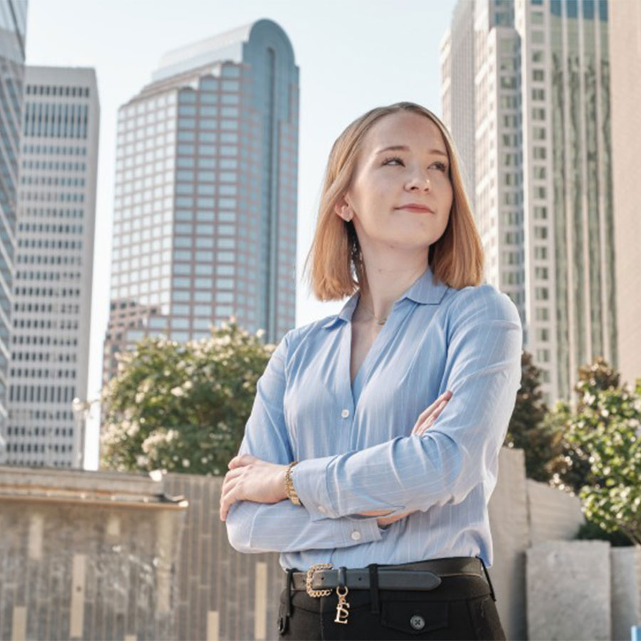 A young adult stands, arms crossed, in a light blue blouse and black slacks in a city park, surrounded by gleaming skyscrapers.