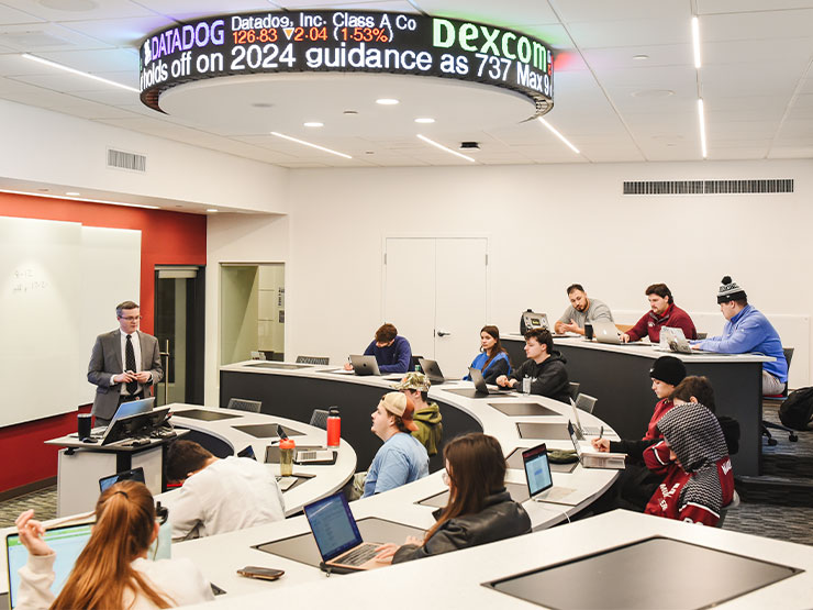 A classroom of students sits below a round stock market ticker on the ceiling.