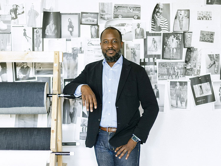 An adult in black blazer and jeans stands in an art studio with black and white photographs covering the wall.
