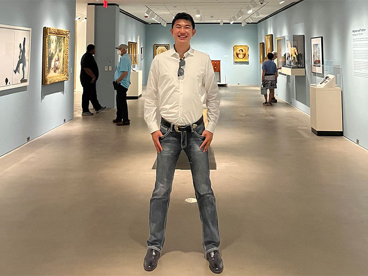 An young adult stands, smiling wide with hands in pockets, in an art museum gallery space.