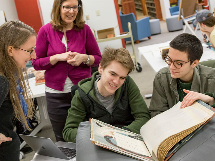 A cluster of students and instructors crowd around and examine an old book of art with yellowed pages.
