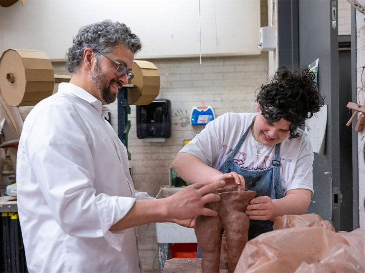 An instructor helps shape a sculpture along side a smiling studio in an art studio.