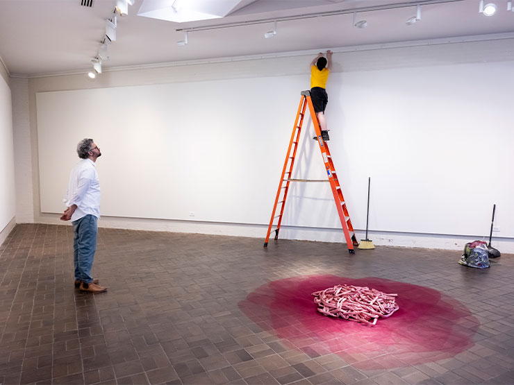 An instructor looks on as a student sets up an art display in a gallery, standing atop an orange ladder against a white wall.