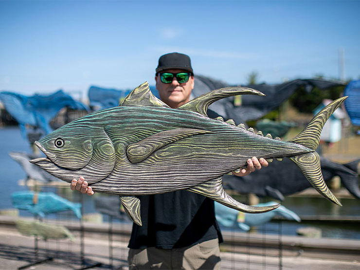 An adult in a baseball cap and sunglasses stands on a dock, holding an intricately carved wooden fish.