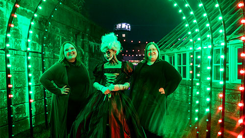 Three adults stand outdoors under neon green lights, one in the middle is wearing dramatic Halloween-themed makeup and dress.