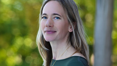 A woman with blonde hair and a green shirt wears a necklace and smiles gently at the camera.