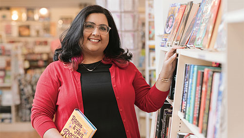A woman in a black shirt and salmon-colored cardigan stands smiling with her hand on a shelf filled with books.