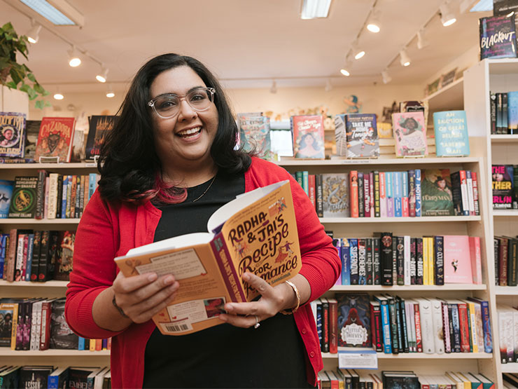 An adult in a black shirt with a salmon-colored cardigan laughs in a bookstore while holding open a book she wrote.
