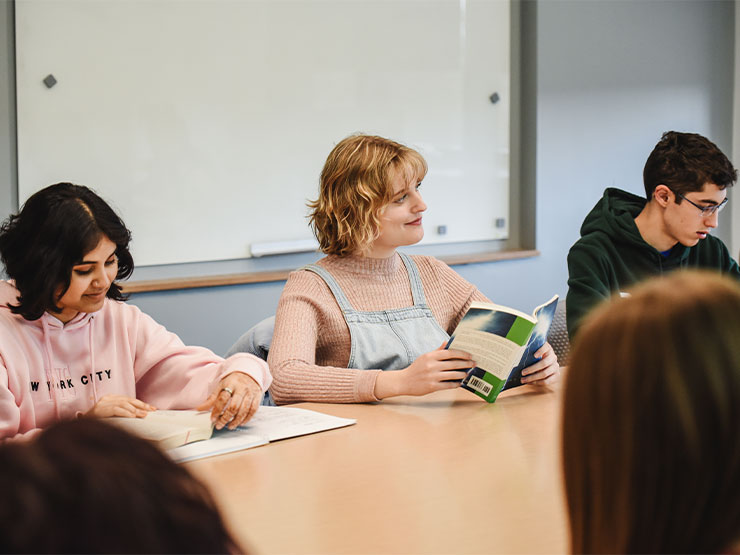 A trio of students sit beside each other at a shared desk space with books open.