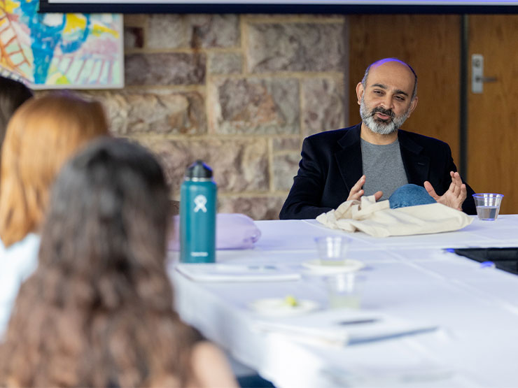 An adult author sits at a table in a classroom and speaks with a group of students.