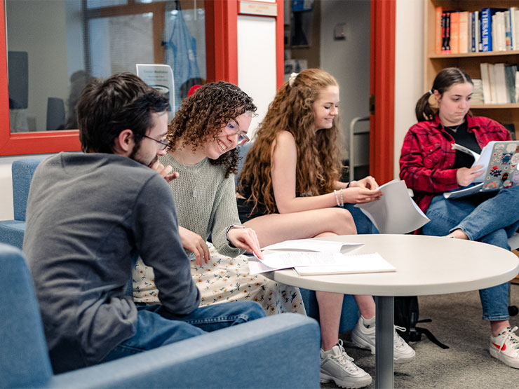 Four students sit around a table in a library room, leafing through packets of papers together.