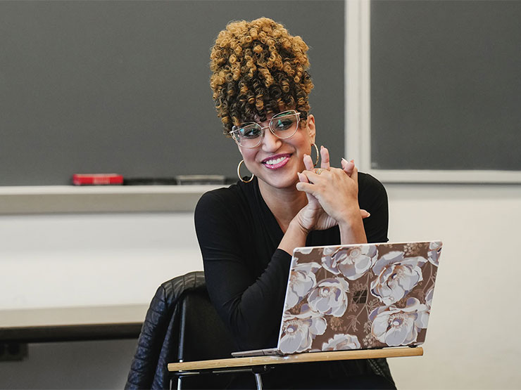 Muhlenberg instructor Emanuela Kucik smiles at a desk with an open laptop in the classroom.