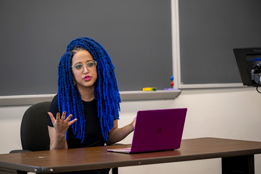 Faculty member seated lecturing at desk positioned in front of chalkboard inside classroom setting.