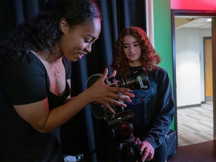 One student adjusts camera equipment with her hands while another looks on in a low-lit studio classroom.