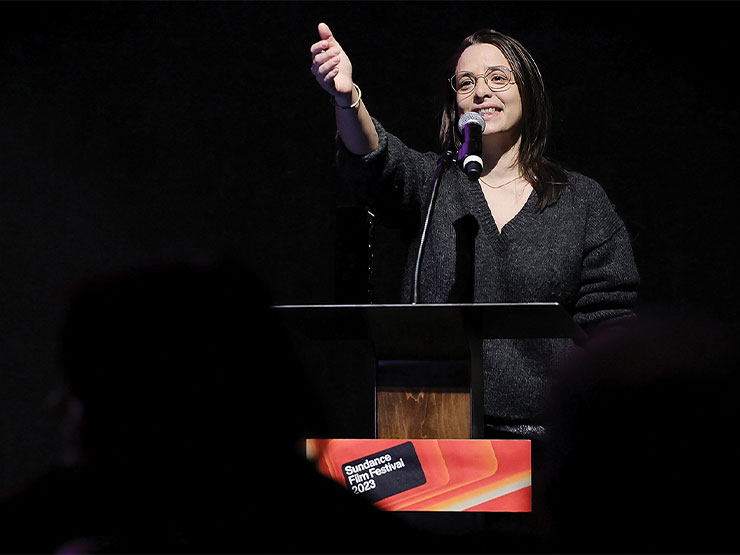 A young adult lit by a spotlight speaks at a podium that reads
