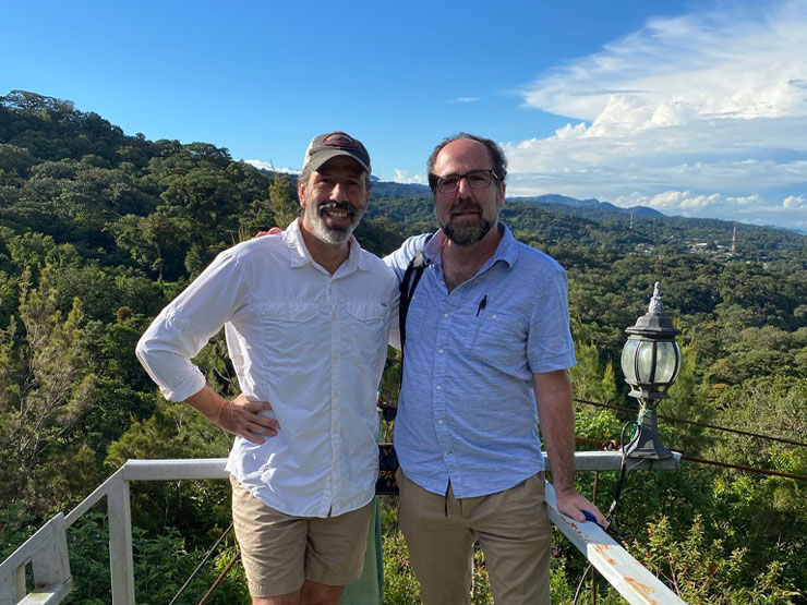 A pair of men stand together on an overlook in a tropical locale under blue skies.