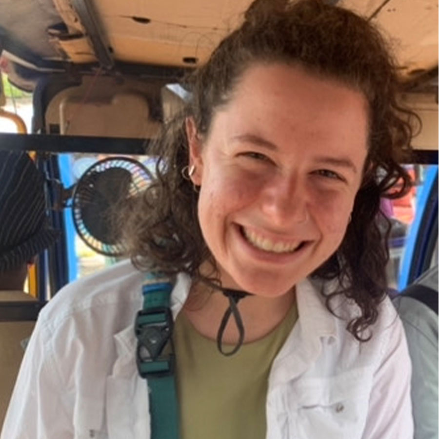 A white young adult with shoulder length brown curly hair smiles to the camera while riding inside a rickshaw.