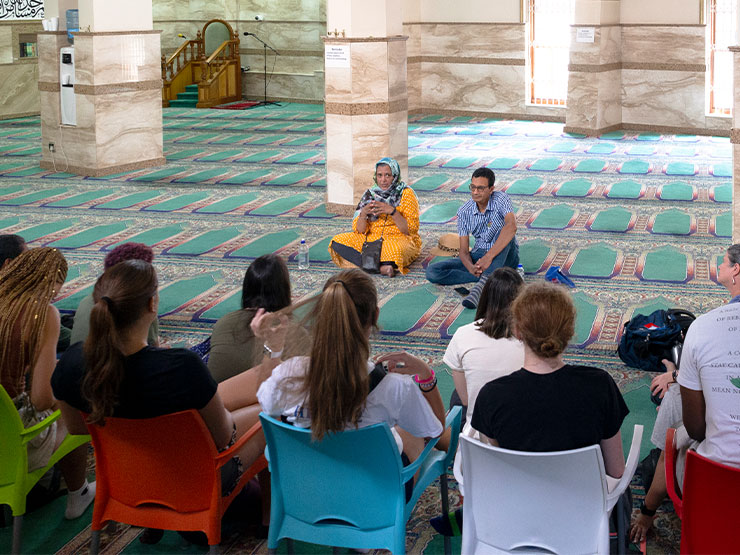 A group of students sit in colorful chairs facing two individuals, one wearing a head scarf, who sit on the floor while speaking in a with colorful carpet and marble walls.